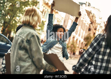 Se sentir heureux. Cheerful young afro american woman holding blank sign board sur la tête et de rire tout en se tenant sur la route autour d'autres militantes. Les droits de l'homme. Concept de protestation Banque D'Images