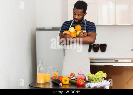 Surpris beau jeune homme portant un teint foncé polo à rayures t-shirt chemise et tablier, debout dans la cuisine blanche, tenue bio frais, les oranges mûres Banque D'Images