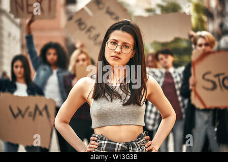 Lutter pour mes droits. Jeune femme confiante dans eyewear en gardant les bras sur les hanches et à la recherche à la caméra au cours d'une manifestation avec groupe de femmes manifestants sur la route. Protestant contre l'extérieur du groupe. Marche des femmes Banque D'Images