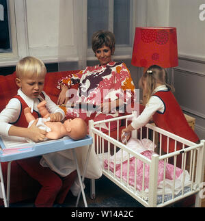 1960 Mère. Une jeune femme est en train de lire un livre pendant que ses enfants jouent ensemble avec des poupées. Le garçon joue avec une poupée et met des vêtements sur elle. Les années 60 ont été la décennie de l'égalité. Suède 1964 Banque D'Images