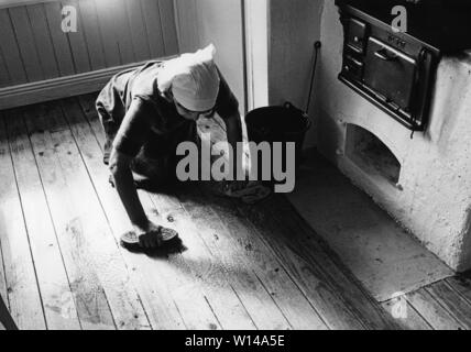 Nettoyage du marbre à l'époque. Une vieille femme est étendue sur le sol et l'utilisation d'une brosse à récurer pour nettoyer le sol de la cuisine. Une vieille cuisinière cuisine en bois est vu. Banque D'Images