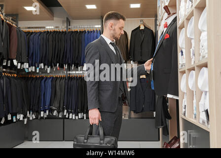 Jeune homme en costume rayé élégant avec beg debout près de mannequin en boutique, le choix, à la toucher et de la veste. De nombreuses étagères en chemise blanche. Banque D'Images