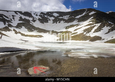 Low angle view of la larme partiellement le lac gelé et recouvert de neige, de montagnes de Rila impressionnant et flèche rouge sur un rocher comme un sentier mark Banque D'Images