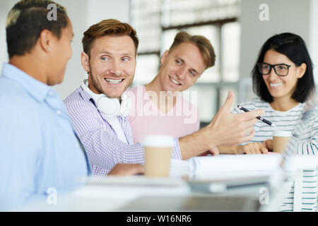 Jeune homme dynamique positive au casque cou assis à table et des gestes main tout en expliquant l'idée de projet à des collègues Banque D'Images