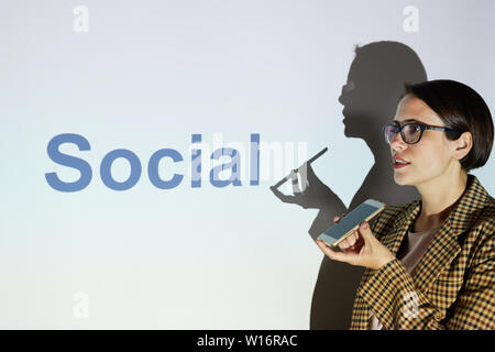 Confiant dans la veste à carreaux attractive businesswoman standing against projecteur avec word Social et de l'enregistrement du son message sur smartphone Banque D'Images