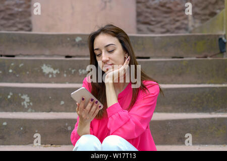 Jeune femme assise sur le ciment extérieur étapes en utilisant son téléphone portable absorbé par la lecture d'un message Banque D'Images