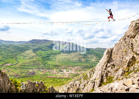 Jeune homme sur une corde tendue au-dessus des falaises déchiquetées. Slackline dans les montagnes avec de belles pistes vertes et des nuages dans l'arrière-plan Banque D'Images