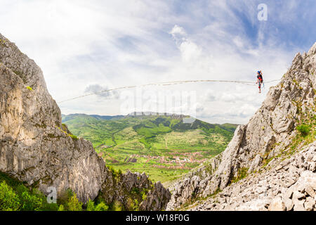 Jeune homme sur une corde tendue au-dessus des falaises déchiquetées. Slackline dans les montagnes avec de belles pistes vertes et des nuages dans l'arrière-plan Banque D'Images