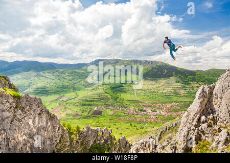 Jeune homme sur une corde tendue au-dessus des falaises déchiquetées. Slackline dans les montagnes avec de belles pistes vertes et des nuages dans l'arrière-plan Banque D'Images