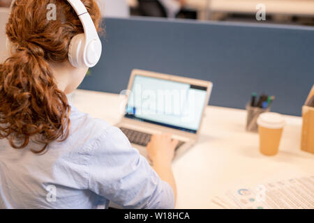 Vue arrière du jeune rousse businesswoman avec queue de l'écoute de la musique dans le casque tout en travaillant avec un fichier texte sur ordinateur portable Banque D'Images