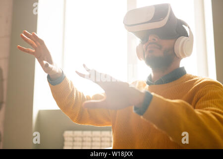 Les jeunes curieux sérieux homme moyen-orientale avec barbe portant un casque sans fil mains des gestes tout en jouant au jeu vidéo lunettes VR Banque D'Images