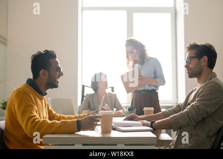 Les jeunes positives des experts en marketing multi-ethnique assis à table dans la salle de réunions et l'échange d'idées tout en discutant nouvelle campagne de publicité Banque D'Images