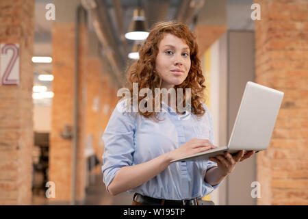 Content confiant jeune femme avec les cheveux bouclés en rouge loft espace de coworking et using laptop while looking at camera Banque D'Images