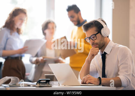 Homme sérieux introspectif manager dans les écouteurs assis à table et lire en ligne le document sur l'ordinateur portable Banque D'Images