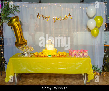 Une table avec un gâteau glacé, cupcakes, cake pops, des cadeaux en face d'une toile blanche décorée avec des ballons gonflés à l'hélium en blanc jaune Banque D'Images