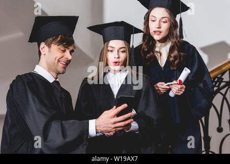 Surpris in graduation caps looking at smartphone près de bel homme Banque D'Images