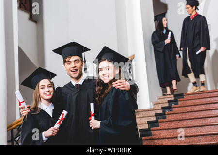 Selective focus of cheerful man in graduation cap hugging girls tout en maintenant les étudiants de diplôme Banque D'Images