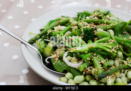 Cuillère à servir dans une salade verte de printemps broccolini, fèves, les échalotes et les haricots verts, parsemés de graines de sésame et de nigelle Banque D'Images