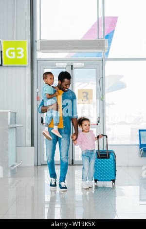 Smiling african american father marcher avec des enfants et des bagages le long de l'attente de l'aéroport de hall Banque D'Images