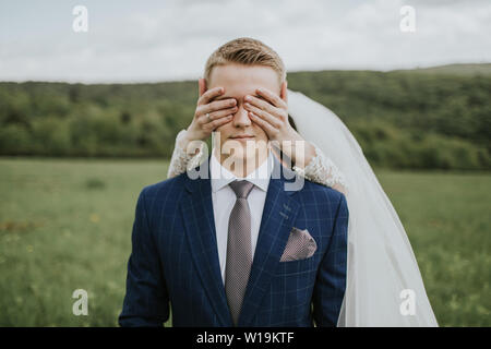 Les yeux du marié mariée couvrant. Portrait de jeune marié en attente sur prairie et bride standing derrière lui, couvrant ses yeux. Banque D'Images