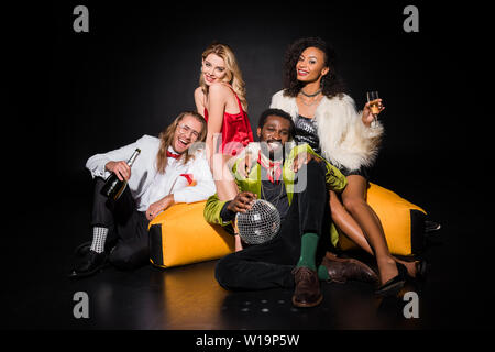 Happy african american man holding dicso boule tout assis près de chaise de sac d'haricot et d'amis sur noir Banque D'Images