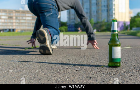 Une bouteille de bière se dresse sur le chemin d'asphalte sur l'arrière-plan des pieds d'un homme qui est de la préparation de l'exécution de sprint Banque D'Images