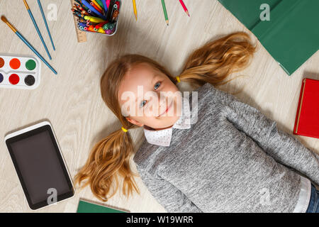 Cute little girl est étendue sur le sol. Un enfant est entouré par un livre, une tablette, peintures, pinceaux, crayons. Banque D'Images