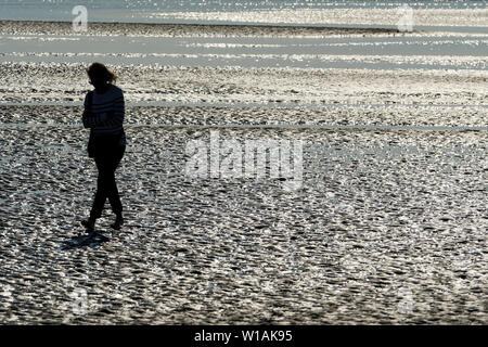 Femme seule vu dans le rétro-éclairage comme elle marche sur le sable humide à marée basse, Le Crotoy, Baie de Somme, Somme, hauts de France, France Banque D'Images