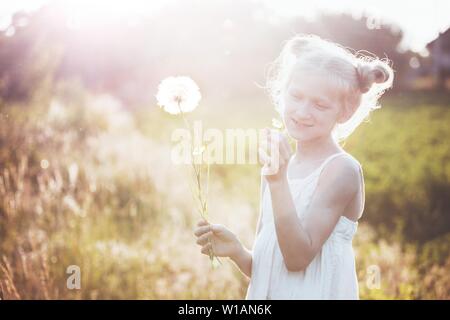 Petite fille blonde holding dandelion et en soufflant sur le coucher du soleil. vacances d'été et de l'humeur Banque D'Images