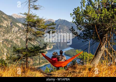 Paire assis dans un hamac avec vue panoramique sur les montagnes et le lac, le lac Plansee, Alpes Ammergau, district de Reutte, Tyrol, Autriche Banque D'Images