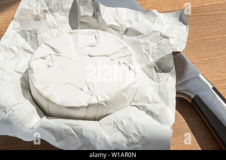 Ouvrez l'emballage du papier de tête entière de camembert et un couteau sous un soleil de la fenêtre. Fromage doux recouvert de moisissure blanche comestible Banque D'Images