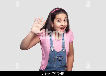 Bonjour. Portrait de surpris belle brune jeune fille en rose occasionnels t-shirt et salopette bleue debout avec étonnement le visage et en agitant sa main à l'intérieur. Banque D'Images