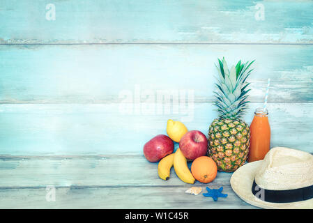 Concept d'été stillife - un tas de fruits frais, chapeau de paille et une bouteille de jus multivitaminé devant un fond bleu en bois rustique. Banque D'Images