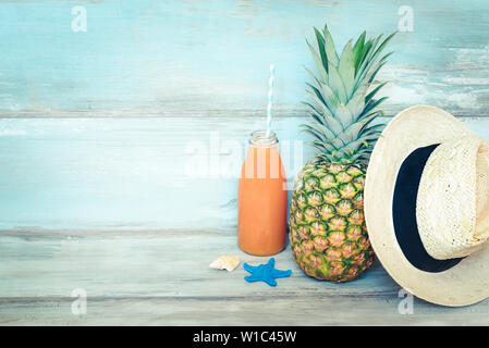 Concept d'été - Ananas mûrs stillife, chapeau de paille et une bouteille de jus multivitaminé devant un fond bleu en bois rustique. Banque D'Images