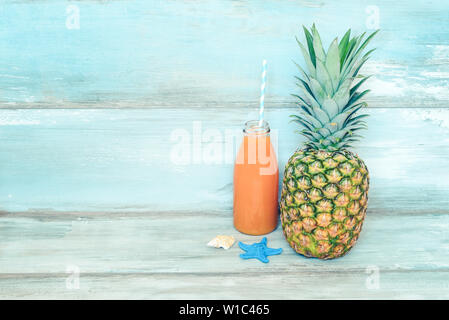 Concept d'été stillife - venu d'ananas et une bouteille de jus multivitaminé devant un fond bleu en bois rustique. Banque D'Images