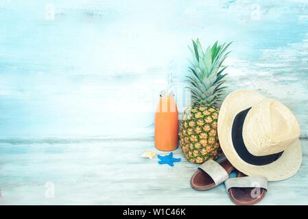 Concept d'été - Ananas mûrs stillife, chapeau de paille, des tongs et une bouteille de jus multivitaminé devant un fond bleu en bois rustique. Banque D'Images