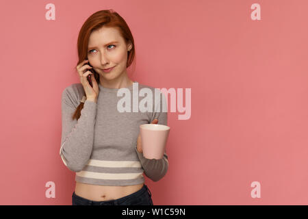 Fille rousse avec une tasse à l'écoute de quelqu'un sur son smartphone avec drôle de visage expression Banque D'Images