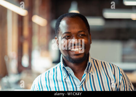 Gai homme à la peau sombre. Cheerful dark-skinned woman smiling chemise rayée alors que se sentir bien au travail Banque D'Images