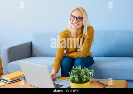 Happy young woman with glasses using laptop at home sitting on couch Banque D'Images