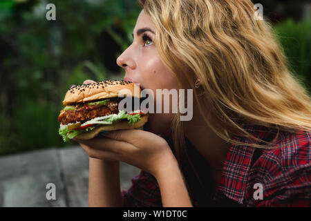 Consommer des étudiants de la restauration rapide. Fille bouchée de très gros burger dans le jardin Banque D'Images