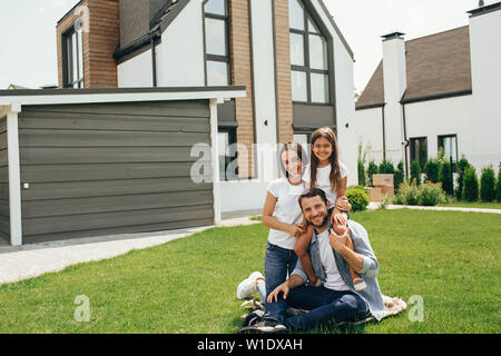 Heteroseksual happy family sitting on grass près de leur nouvelle maison. Famille assis sur la pelouse, l'achat de nouveaux big home . Banque D'Images