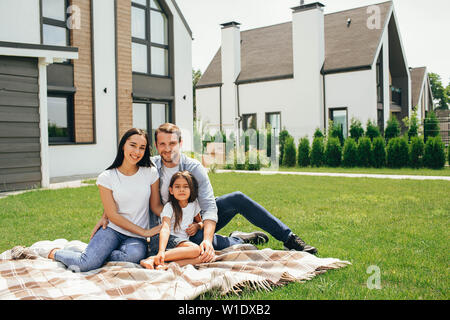 Heteroseksual happy family sitting on grass près de leur nouvelle maison. Famille assis sur la pelouse, l'achat de nouveaux big home . Banque D'Images