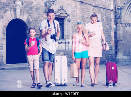 Jeune famille de quatre avec des valises à marcher le long en utilisant un téléphone Banque D'Images