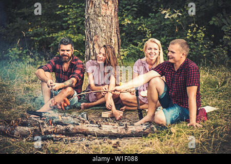 Deux couples camping dans les bois. Saucisses à frire les gars sur le feu. Smiling friends sitting autour de feu de camp le soir Banque D'Images