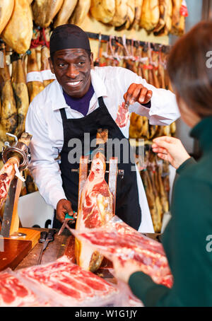 Smiling African American worker au compteur dans une boucherie, la vente à la clientèle féminine ibérique jamon Banque D'Images