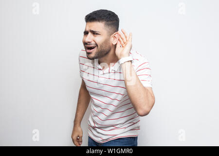 Quoi ? Je ne peux pas vous entendre. Portrait de jeune homme barbu beau attentif en t-shirt à rayures debout avec la main sur l'oreille et veulent entendre quelque chose. en Banque D'Images