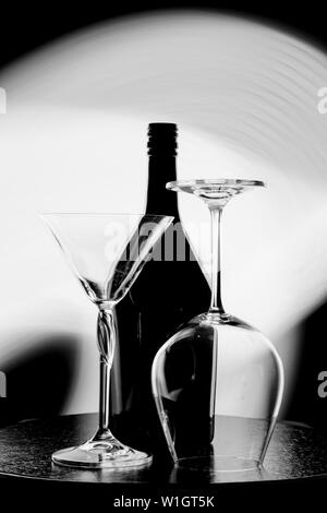 Une photo en noir et blanc d'une bouteille de vin et deux verres contre le point lumineux sur le mur noir Banque D'Images