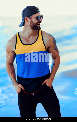 L'homme tatoué barbu au complexe sportif dans un T-shirt, chapeau et lunettes de soleil sur le fond de la mer. Banque D'Images