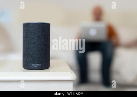 Une 2e génération de l'écho et le président d'Amazon smart Personal assistant est vu dans un salon dans une maison alors qu'un homme utilise un ordinateur portable dans l'arrière-plan. Banque D'Images