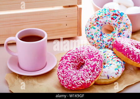 Donuts colorés sur papier rose avec tasse de café, les guimauves et une caisse sur table. Vue en gros plan. Banque D'Images